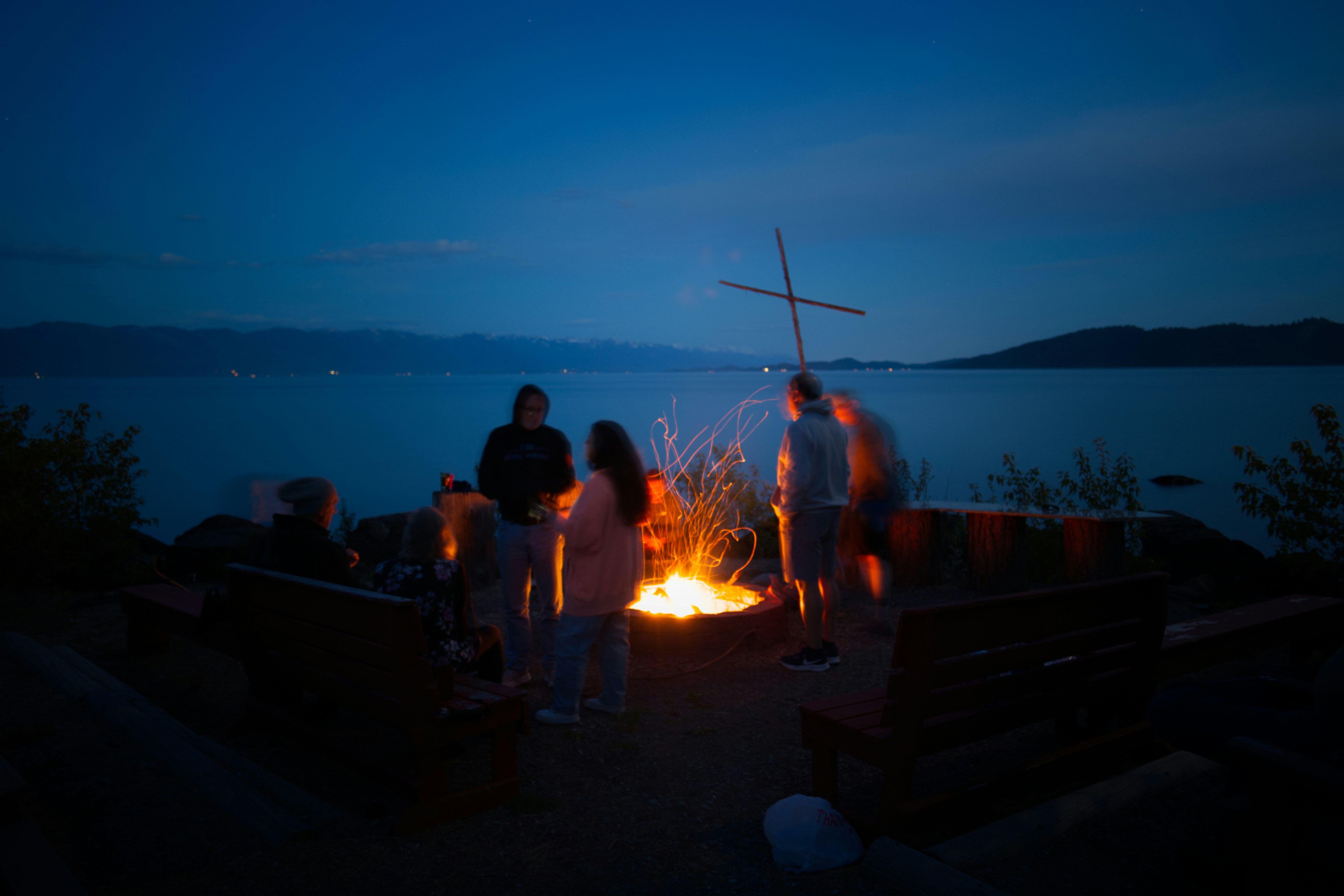 Gruppe von Menschen um ein Lagerfeuer bei Nacht am See, im Hintergrund ein Holzkreuz und Berge.