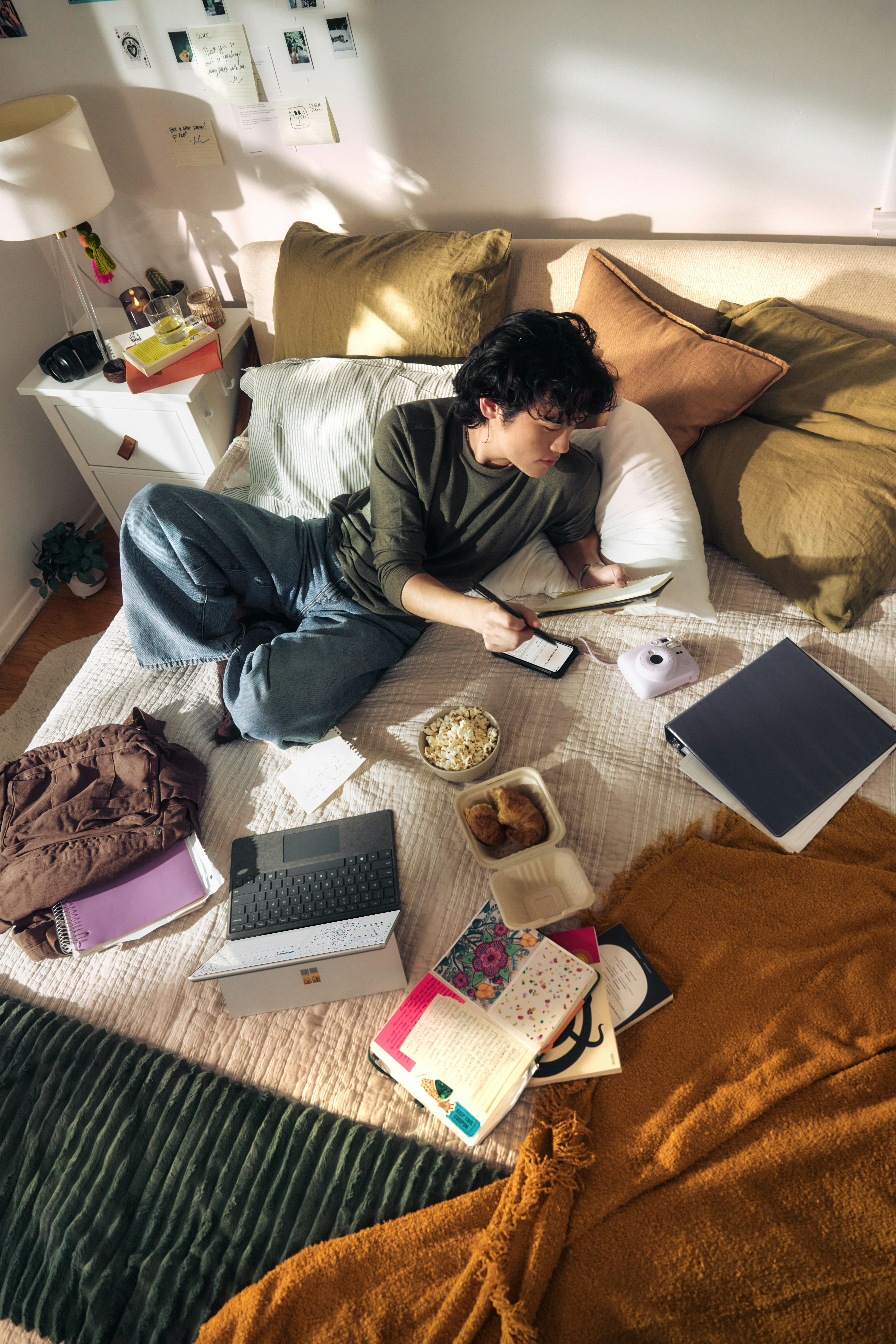 Person sitzt auf dem Bett umgeben von Laptop, Büchern, Snacks und Tablet und schreibt in ein Notizbuch.