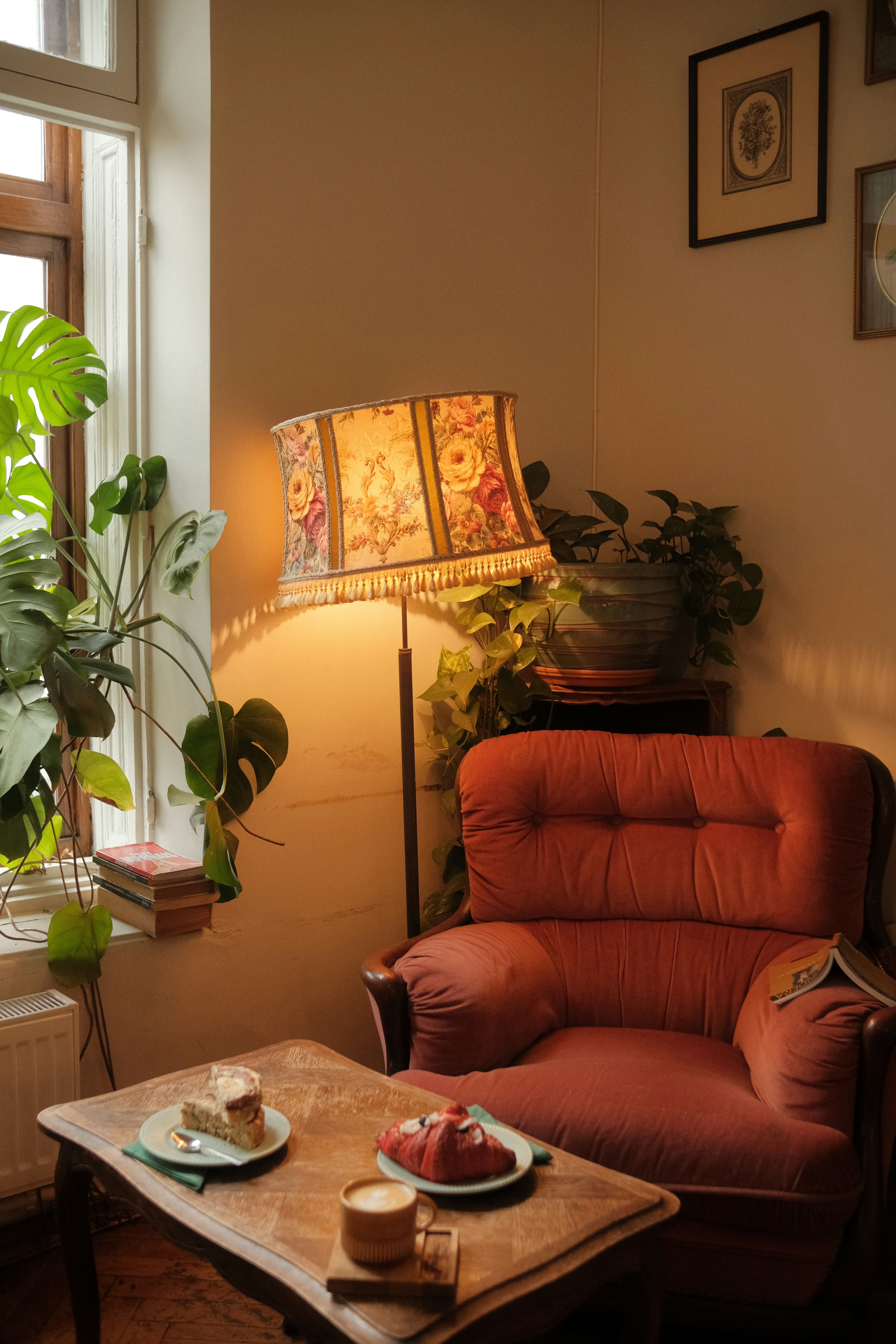 Gemütliche Leseecke mit rotem Sessel, Stehlampe mit Blumenschirm, Pflanzen und Kuchenteller auf Couchtisch.